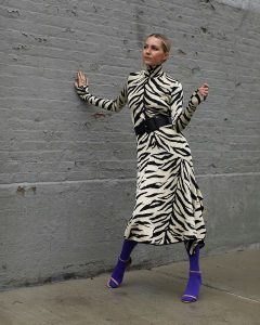 Stylowa kobieta w zebra sukience i niebieskich butach na tle szarej ściany.